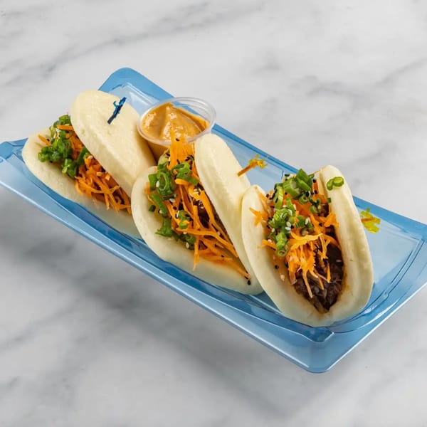 Four Bun Buds bao buns with meat, shredded carrots, green onions, and sesame seeds on a blue tray.