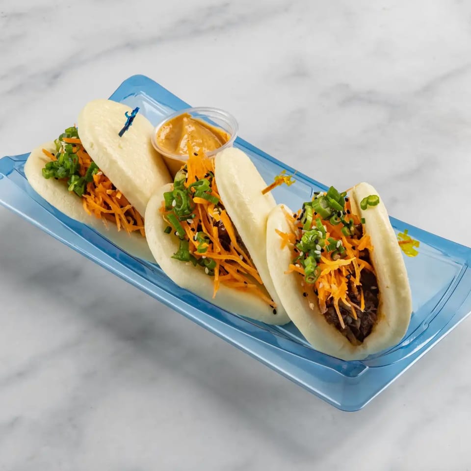 Four Bun Buds bao buns with meat, shredded carrots, green onions, and sesame seeds on a blue tray.